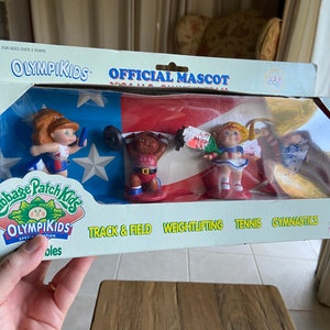 May include: A set of four Cabbage Patch Kids Olympikids dolls, each dressed in a different sport uniform. The dolls are posed in front of an American flag backdrop. The sports represented are track and field, weightlifting, tennis, and gymnastics. The packaging includes the text "OLYMPIKIDS OFFICIAL MASCOT".