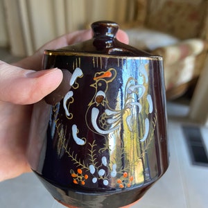 May include: A dark brown ceramic sugar bowl with a lid, featuring a white rooster design with orange accents. The bowl has a glossy finish and gold trim.