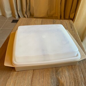May include: A beige plastic food storage container with a clear plastic lid. The lid has a small circle in the center.