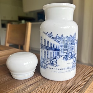 May include: White ceramic jar with a matching lid. The jar features a blue illustration of a European town scene. The jar and lid are sitting on a wooden surface. The jar has a cylindrical shape.