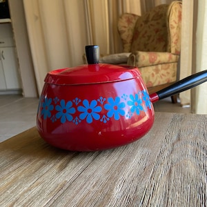 May include: Red enamel fondue pot with a black handle and lid. The pot has a blue and white floral design around the rim.
