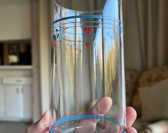 May include: A clear, tall drinking glass with a decorative blue and red floral design. The glass has a blue and red band near the top and bottom. The glass is held in a hand.