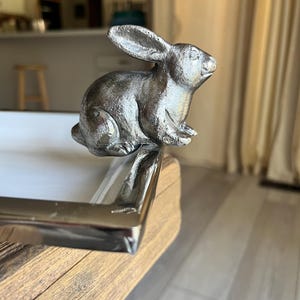 May include: A silver-colored rabbit figurine perched on the corner of a wooden tray with a silver metal trim. The rabbit has detailed fur texture and is in a seated position, with its ears perked up.