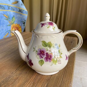 May include: A white teapot with a floral design featuring purple, white, and yellow flowers. The teapot has a gold trim around the lid and spout.