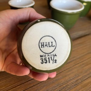 May include: A collection of green ceramic cups with white interiors. The bottom of one cup displays the text "HALL" inside a circle, along with "MADE IN USA" and the number "351 1/2".