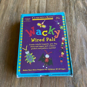 May include: A boxed craft kit labeled "Curiosity Kits Wacky Wired Pals." The box is purple with colorful illustrations of wired creatures. Text on the box says "Create colorful magnets, pins, key chain and zipper pull critters." The box is on a wooden surface.