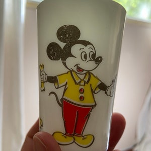 May include: White plastic cup with a cartoon image of Mickey Mouse wearing a yellow shirt and red pants. Mickey is holding a bone in his right hand and a small object in his left hand.