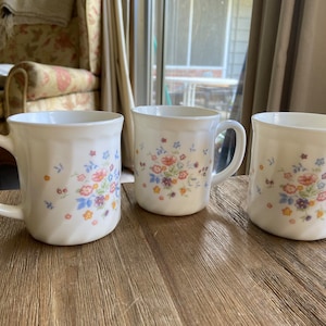 May include: Three white ceramic mugs with a floral pattern in shades of pink, blue, and yellow. The mugs have a rounded shape and a handle on the side.
