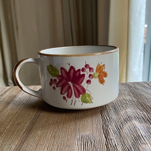 May include: A white ceramic mug with a brown handle and a floral design featuring red, orange, and green flowers.