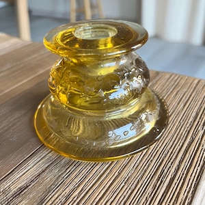 May include: A yellow glass candle holder with a round base and a decorative floral pattern. The candle holder has a cylindrical body with a flared top, and is sitting on a wooden surface.