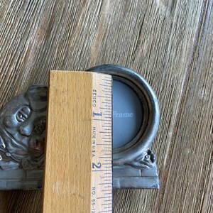 May include: A silver-toned metal picture frame with a round opening and ornate details. The frame is approximately 2 inches wide and 3 inches tall. The frame is sitting on a wooden surface.