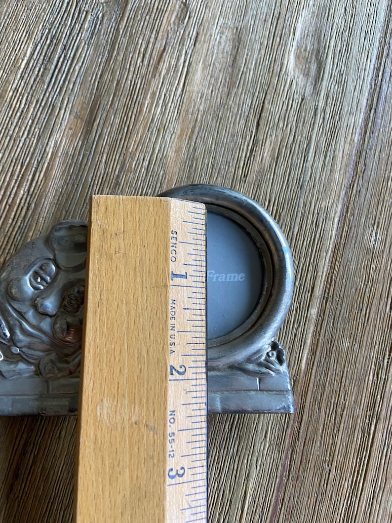 May include: A silver-toned metal picture frame with a round opening and ornate details. The frame is approximately 2 inches wide and 3 inches tall. The frame is sitting on a wooden surface.