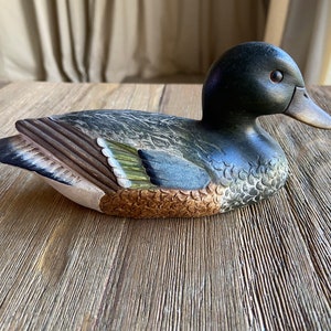May include: A wooden duck decoy with a dark green head, brown and white feathers, and a green and brown wing. The decoy is painted with realistic details and is sitting on a wooden surface.