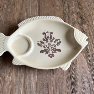 May include: A ceramic fish-shaped serving platter with a brown floral design and a small dip bowl. The platter is beige with a brown rim.