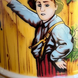 May include: A detailed illustration on a ceramic surface depicts a young person in a blue shirt, red striped pants, and a yellow hat, leaning against a wooden fence. The artwork features a gold trim and a pastoral theme.