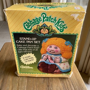 May include: A yellow box with a Cabbage Patch Kids stand-up cake pan set. The box features a picture of a Cabbage Patch Kid doll holding a pink ribbon. The text on the box reads "Cabbage Patch Kids Stand-Up Cake Pan Set".
