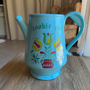 May include: A light blue ceramic cookie jar shaped like a teapot with a floral design and the words "cookie pot" written on the front.