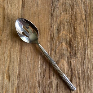 May include: A silver slotted spoon with a bamboo-style handle. The spoon is on a wooden surface.