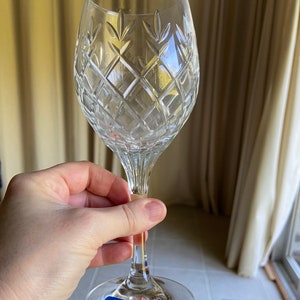 May include: A clear glass wine glass with a cut crystal design. The glass has a long stem and a wide bowl. The glass is sitting on a white tiled surface. The glass has a blue sticker on the bottom that says "ONEIDA SCHOTT ZWIESEL".