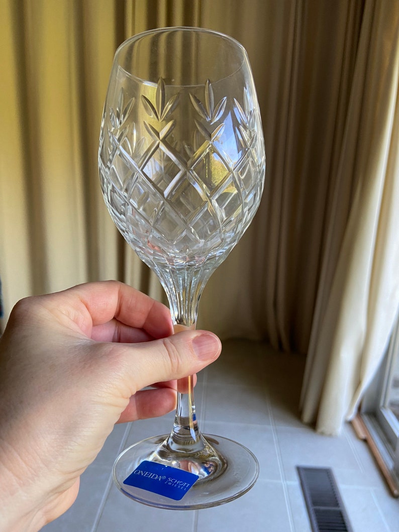May include: A clear glass wine glass with a cut crystal design. The glass has a long stem and a wide bowl. The glass is sitting on a white tiled surface. The glass has a blue sticker on the bottom that says "ONEIDA SCHOTT ZWIESEL".