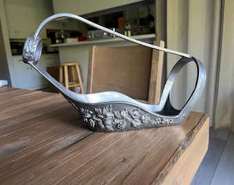 May include: A silver-colored wine bottle holder with a decorative floral and grape design. The holder has a handle and a curved design, suitable for holding a wine bottle. The holder is made of metal.