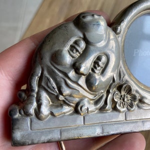 May include: Silver-toned photo frame with a sculpted cherub face and a floral design. The frame has a round opening for a photo and the text "Photo" is printed on the glass.