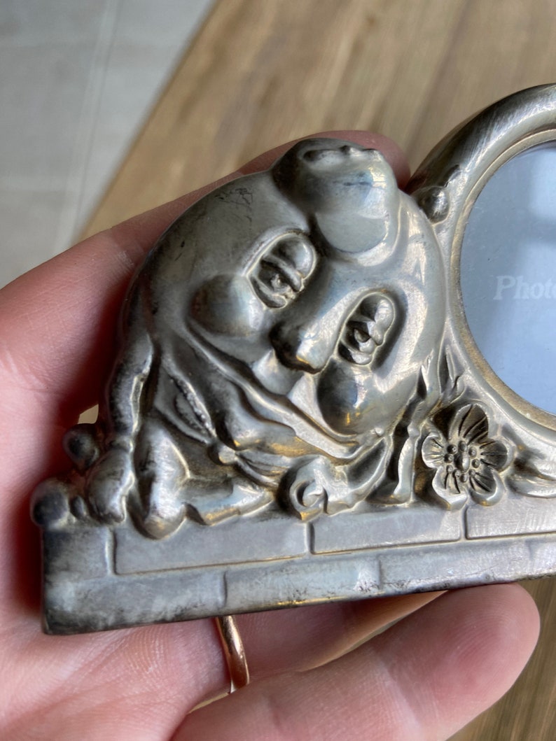 May include: Silver-toned photo frame with a sculpted cherub face and a floral design. The frame has a round opening for a photo and the text "Photo" is printed on the glass.