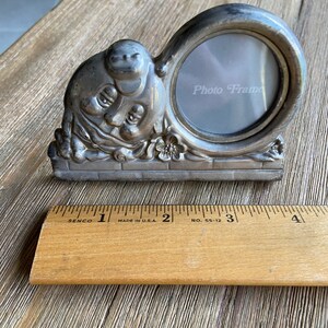 May include: Silver photo frame with a whimsical design featuring a cherubic face and floral accents. The frame has a round opening for a photo and the words "Photo Frame" are printed on the glass.