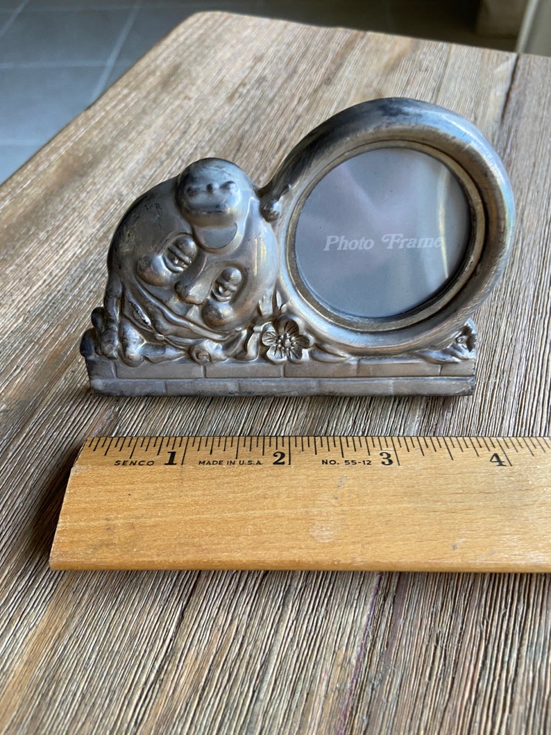 May include: Silver photo frame with a whimsical design featuring a cherubic face and floral accents. The frame has a round opening for a photo and the words "Photo Frame" are printed on the glass.