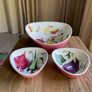 May include: Three ceramic bowls with pink rims and white interiors. Each bowl features a different hand-painted vegetable design. The largest bowl has a design of fall leaves, the medium bowl has a design of a beet, onion, and leek, and the smallest bowl has a design of an eggplant.