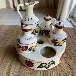 May include: A white ceramic condiment set with a strawberry and leaf design. The set includes a pitcher labeled "OIL", a salt shaker labeled "SALT", a pepper shaker labeled "PEPPER", and a mustard jar labeled "MUSTARD".