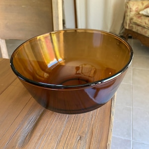 May include: A large, amber-colored glass mixing bowl sits on a wooden surface. The bowl is round and has a smooth, shiny finish.