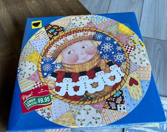 May include: A blue box with a circular illustration featuring a smiling child wearing a hat and scarf, surrounded by snowmen and a patchwork design. The box has a "New! Lower Price!" sticker.