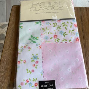 May include: A packaged Fashion Classics oval tablecloth. The tablecloth is divided into four sections with floral patterns in pink, white, and light green. The size is 60x84 inches (152cm x 213cm). The packaging reads "Table Linens by Fashion Industries."