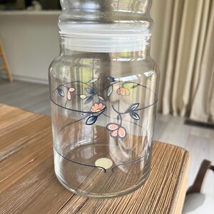 May include: A clear glass jar with a lid, decorated with a floral pattern in pink, blue, and green. The jar is cylindrical and sits on a wooden surface. The lid is a frosted glass.