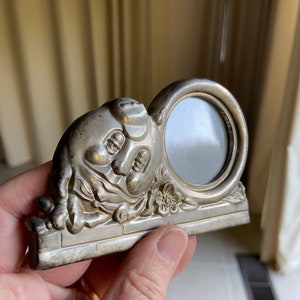 May include: Silver-toned picture frame shaped like a pig with a round opening for a photo. The pig is sitting on a base with a floral design.