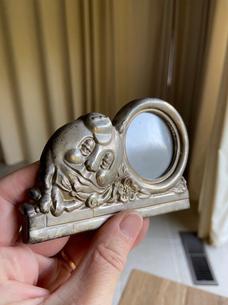 May include: Silver-toned picture frame shaped like a pig with a round opening for a photo. The pig is sitting on a base with a floral design.