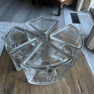 May include: Four clear glass serving dishes with triangular lids. Each dish has a curved side and a handle. The lids have a decorative design in the center. The dishes are arranged on a wooden surface.