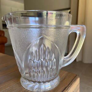 May include: A clear glass pitcher with a textured design. The pitcher has a wide mouth and a handle.
