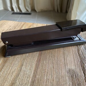 May include: A brown metal stapler with a black top and a silver base. The stapler is sitting on a wooden surface.