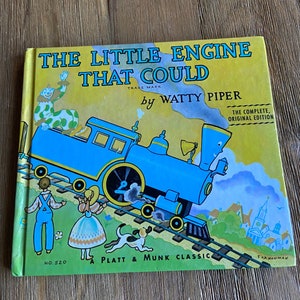 May include: A vintage children's book cover featuring a blue train engine with a smiling face, pulling a train car. The book title is "The Little Engine That Could" by Watty Piper. The cover also features a clown, a girl, a boy, and a dog.