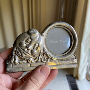 May include: Silver photo frame shaped like a cartoon character with a round picture opening. The frame has a brick base and a flower detail. The text "Photo Frame" is visible in the picture opening.