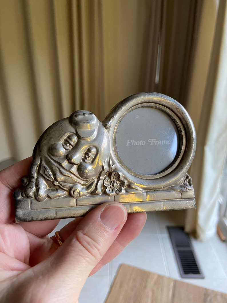 May include: Silver photo frame shaped like a cartoon character with a round picture opening. The frame has a brick base and a flower detail. The text "Photo Frame" is visible in the picture opening.