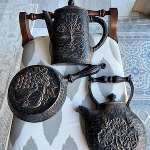 May include: Three antique, dark bronze kitchenware items: a coffee pot, a frying pan, and a kettle. The coffee pot features a floral design, the frying pan has a grape motif, and the kettle showcases a floral pattern. All pieces have textured surfaces.