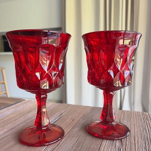 May include: Two ruby red crystal wine glasses with a detailed, cut-glass design. The glasses have a tulip-shaped bowl, a slender stem, and a circular base. They are placed on a wooden surface, showcasing their vibrant color and intricate patterns.