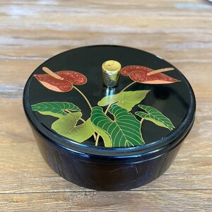 May include: A black round trinket box with a gold knob on the lid. The lid is decorated with a floral design of green leaves and red flowers.