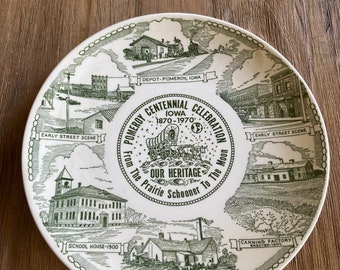 May include: A white ceramic plate with a green border and illustrations of historical scenes from Pomeroy, Iowa. The plate commemorates the town's centennial celebration from 1870 to 1970. The text reads "Pomeroy Centennial Celebration, Iowa 1870-1970, From the Prairie Schooner to the Moon, Our Heritage".