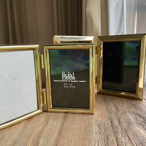 May include: Three gold-toned picture frames with a hinged design. The frames are made of solid brass and are hand polished and lacquered. The middle frame has the text "Solid Brass" and "hand polished & lacquer coated" and the dimensions "3 1/2" x 5" / 9cm x 13cm".