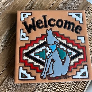 May include: Square ceramic tile with the word "Welcome" in black, featuring a gray wolf illustration and a Southwestern-inspired geometric design in red, white, and teal. The tile has a tan background and a brown border.