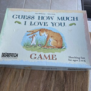 May include: A board game box with a light blue background featuring a cartoon illustration of two rabbits. The box has the title "Guess How Much I Love You" and the text "Game" in large, red letters. The box is designed for ages 3 to 6.